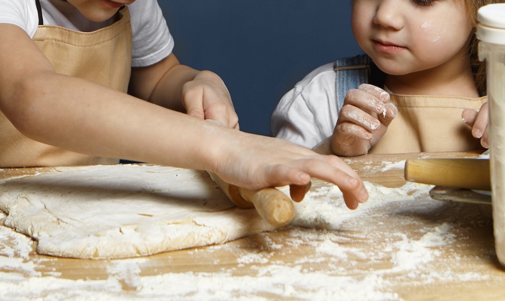 Adorable siblings boy and girl baking cookies together, standing at kitchen table with bottle of milk, flour, flattening dough using rolling pin. Family, childhood, homemade bakery, joy and happiness